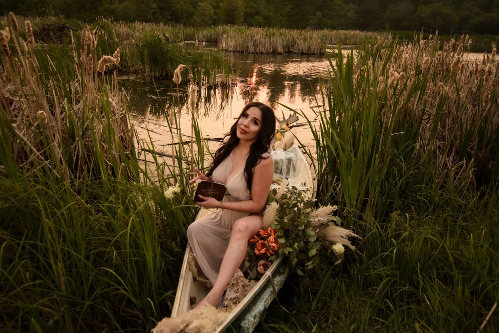 A person lounges in a wooden boat surrounded by tall reeds and wildflowers at sunset on a tranquil pond.