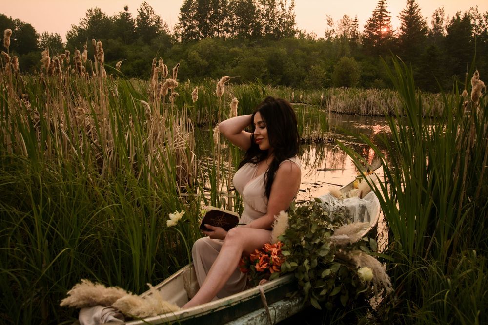 A serene lake view at dusk with a woman in a white dress posing gracefully in a rustic boat among cattails.