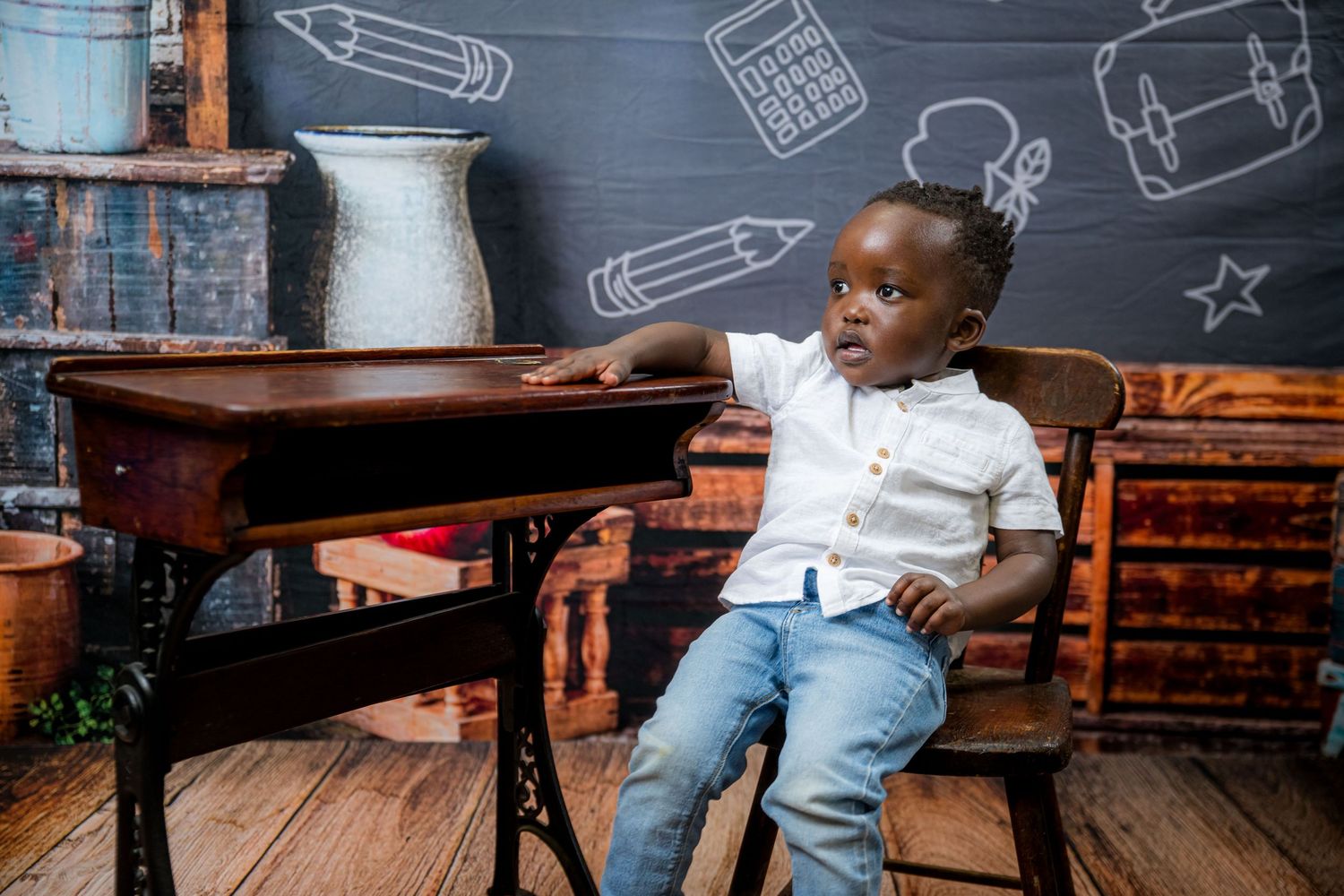 Young student sits at a classroom desk with chalkboard drawings of school supplies on the wall behind them.