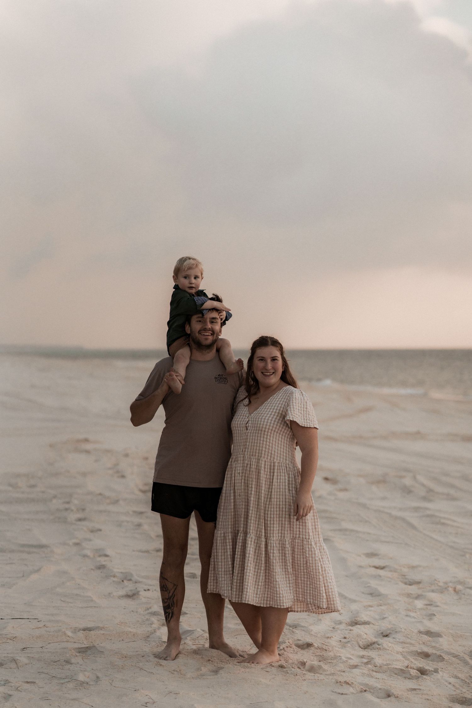 A family poses together barefoot on a misty beach during sunset, wearing casual attire.