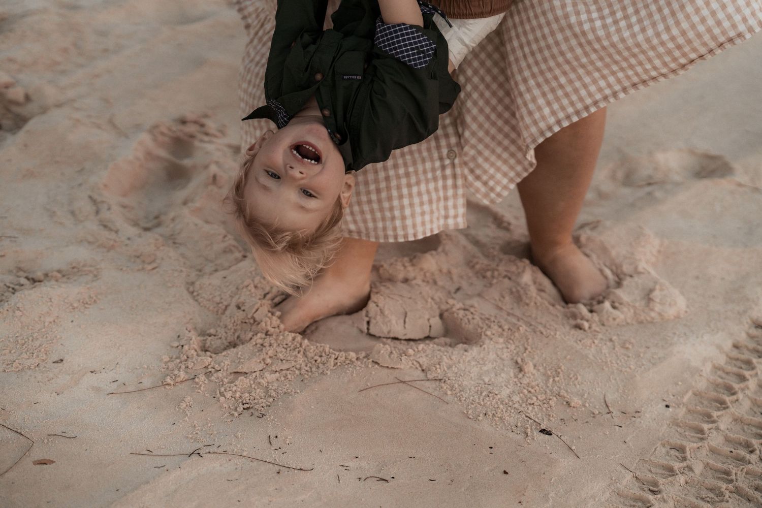 A child lies playfully in the sand at the beach while someone in a checkered dress stands nearby.