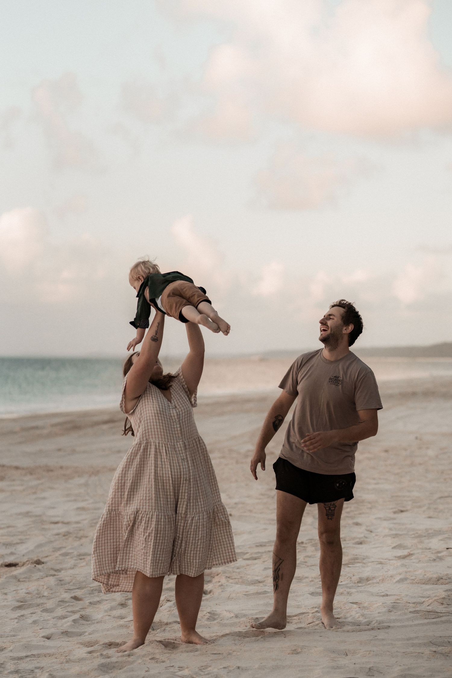 A couple in coordinating beige outfits poses playfully on a sandy beach at sunset taking selfies together.