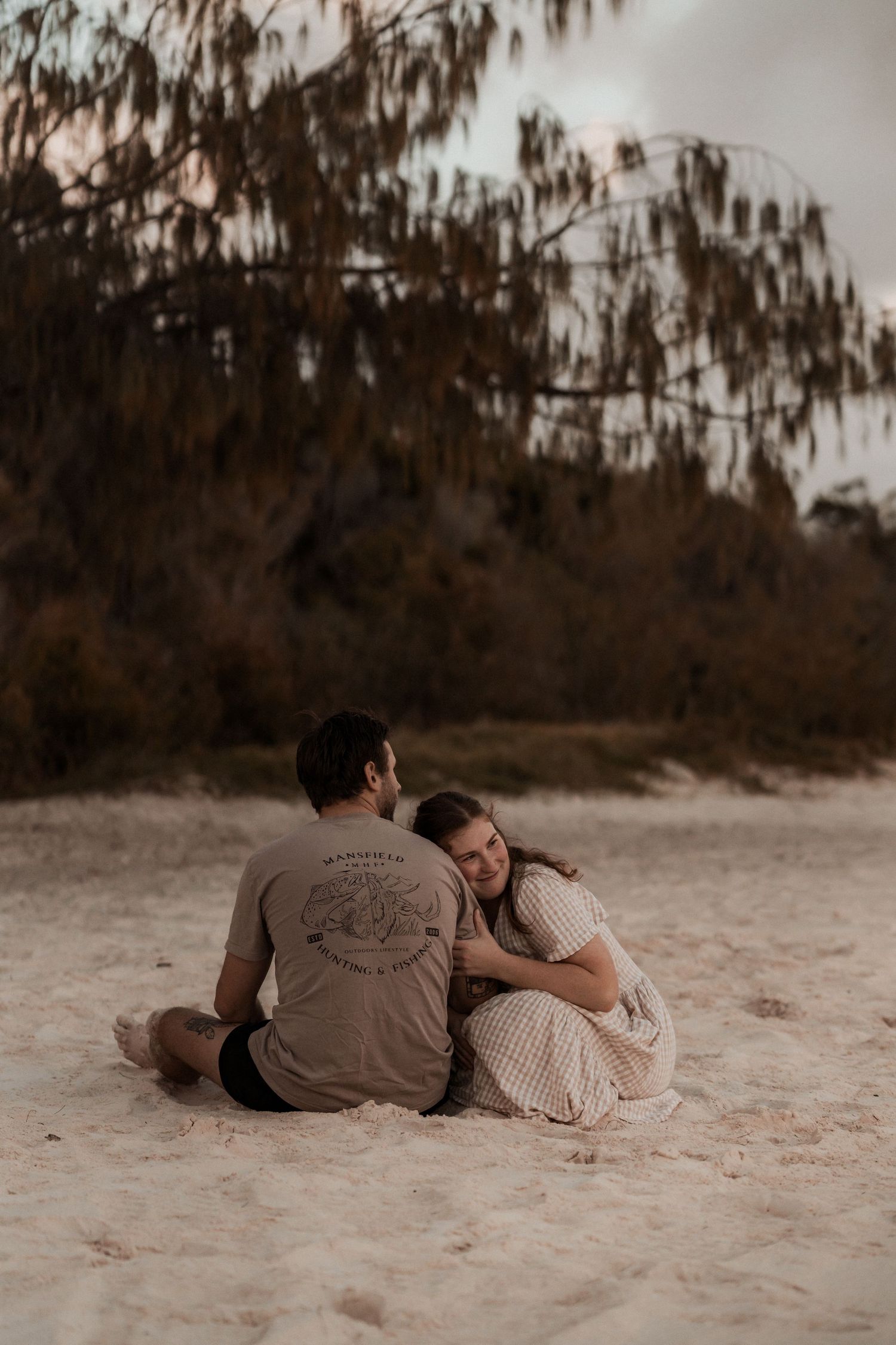A couple sits close together on a sandy beach at sunset with Spanish moss hanging from trees in the background.