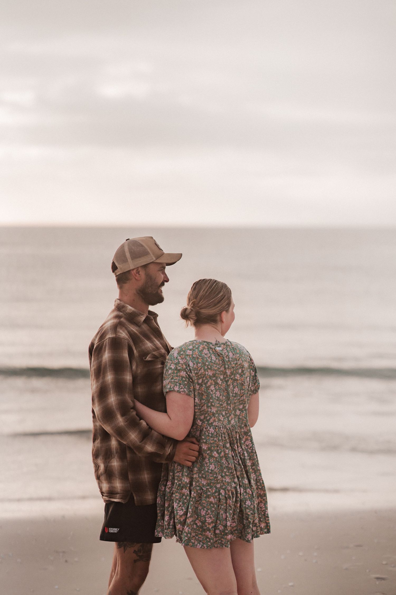 A couple embraces while standing on a misty beach at sunset wearing casual attire.