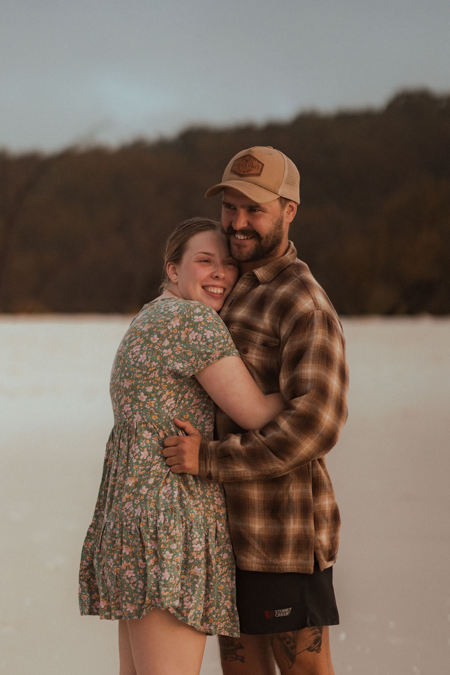 A couple embraces in casual clothing on a beach during golden hour, creating a romantic outdoor scene.