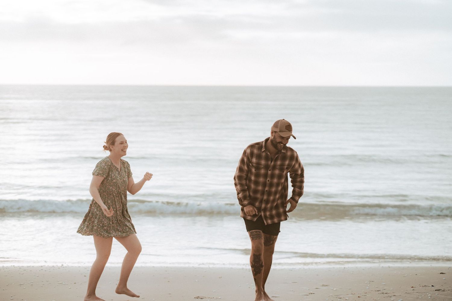Two people playfully run and chase each other along a misty beach shoreline at twilight.