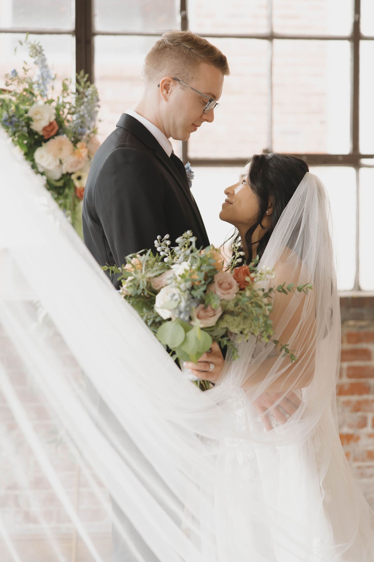 Newlywed couple shares intimate moment in front of window with flowing veil and floral arrangements.
