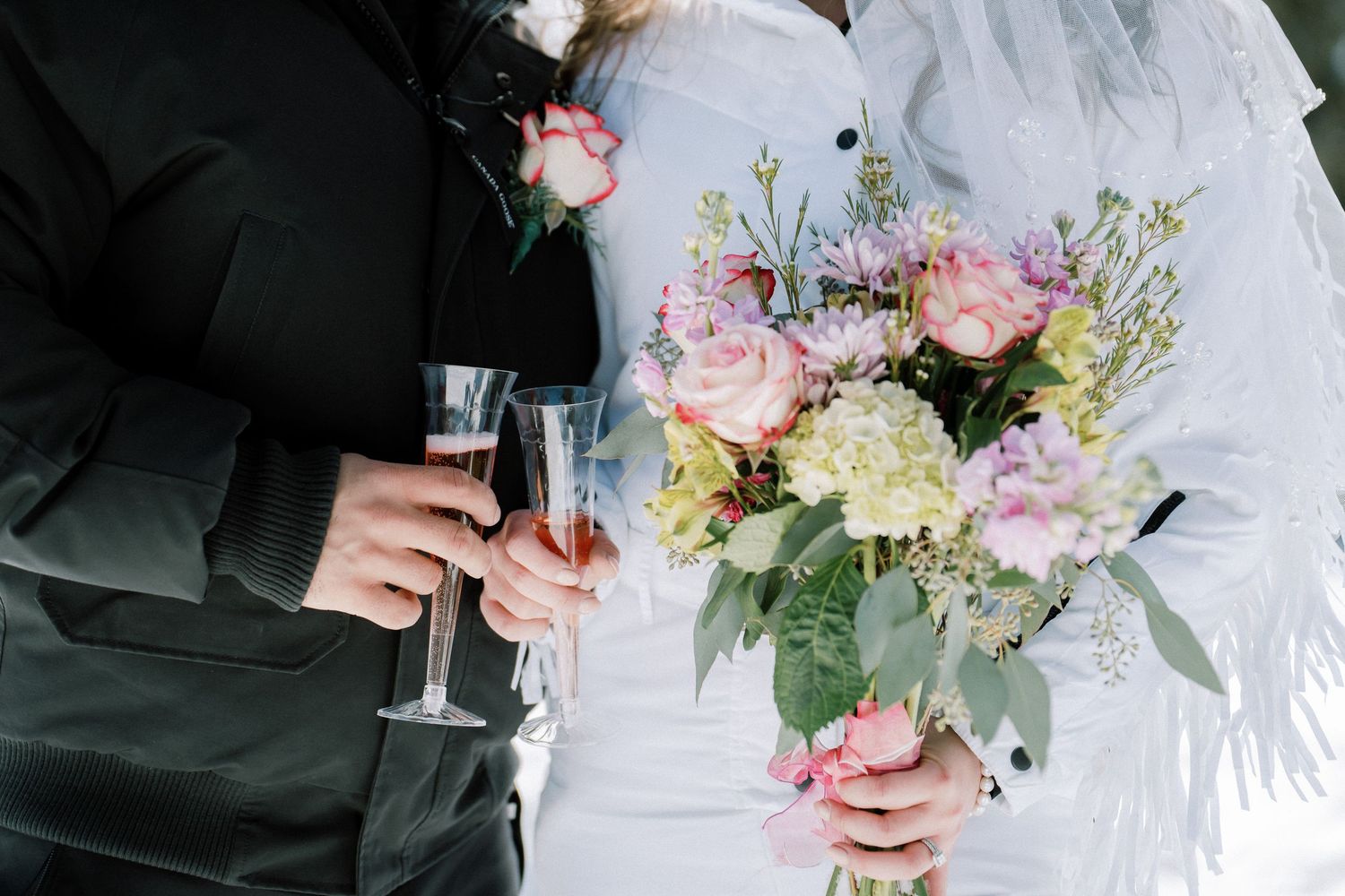 Bride in white dress holds pink bouquet while clinking champagne glass with groom in black suit.
