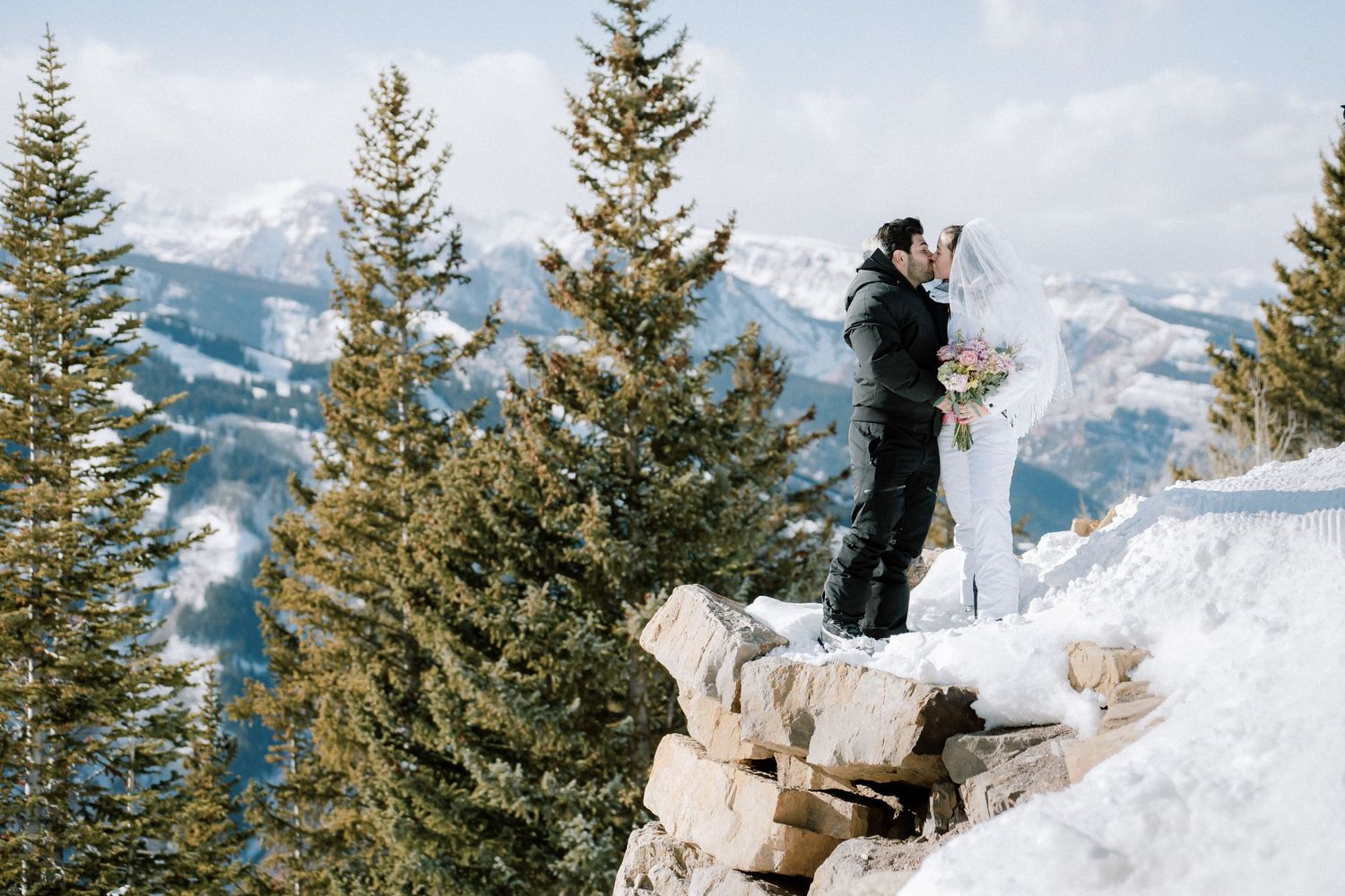 Skier standing on snowy mountain peak overlooking winter landscape with evergreen trees and distant peaks.