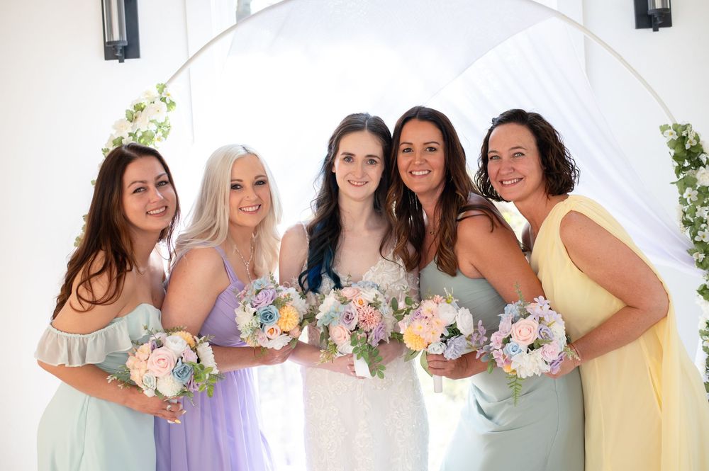 Bridal party poses together in pastel colored dresses holding floral bouquets under a white arch.