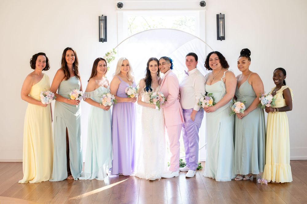 Wedding party in pastel floor-length dresses stands together against a neutral wall holding delicate bouquets.