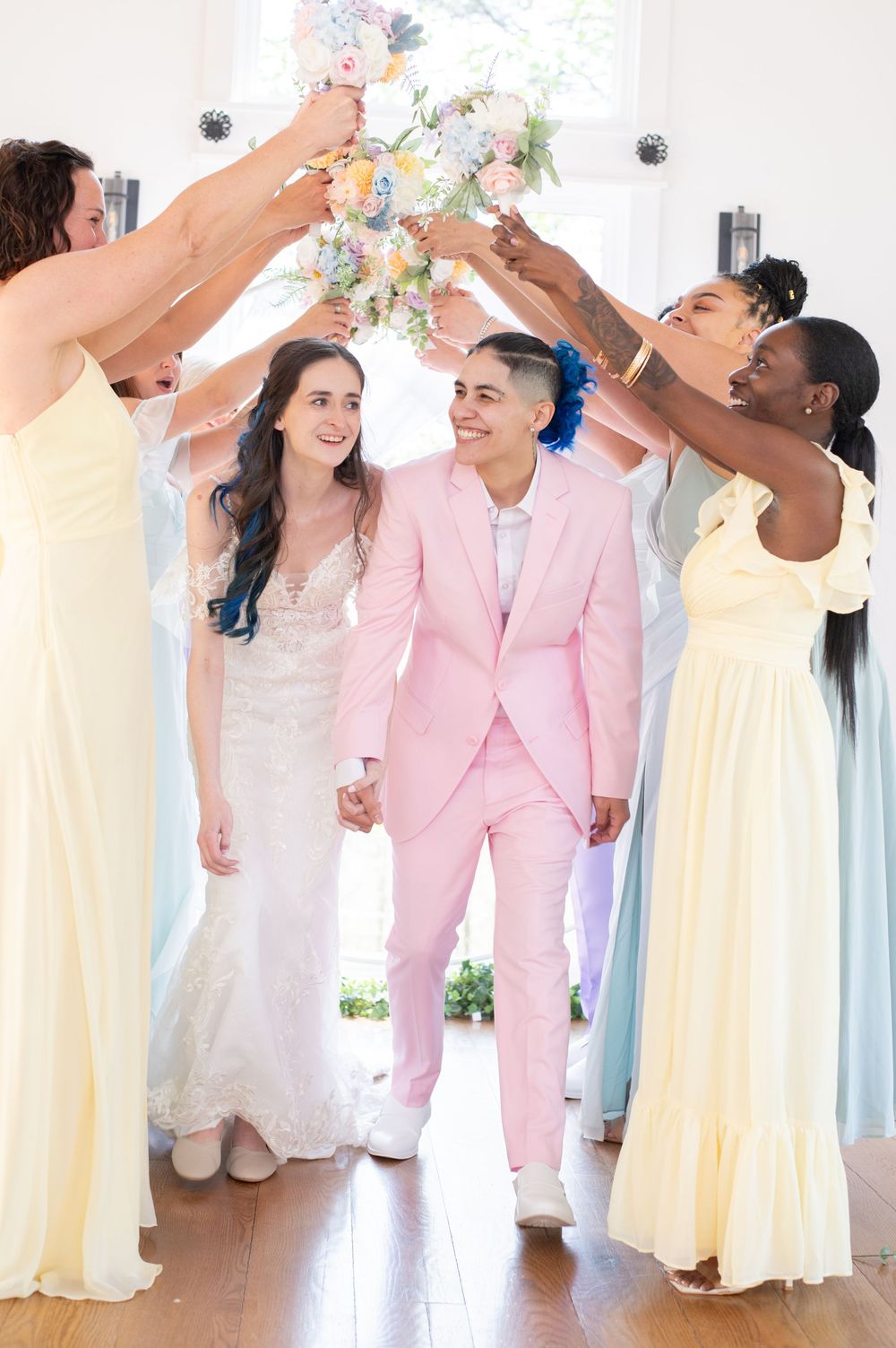 Wedding party wearing yellow dresses and pink suit celebrate joyfully by raising their glasses in a sunlit venue.