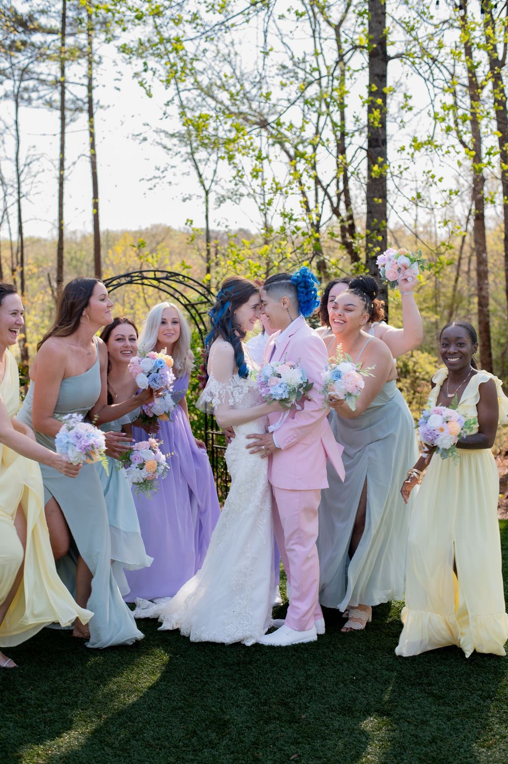 Wedding party members in pastel dresses celebrate outdoors with bouquets beneath a floral arch during spring ceremony.
