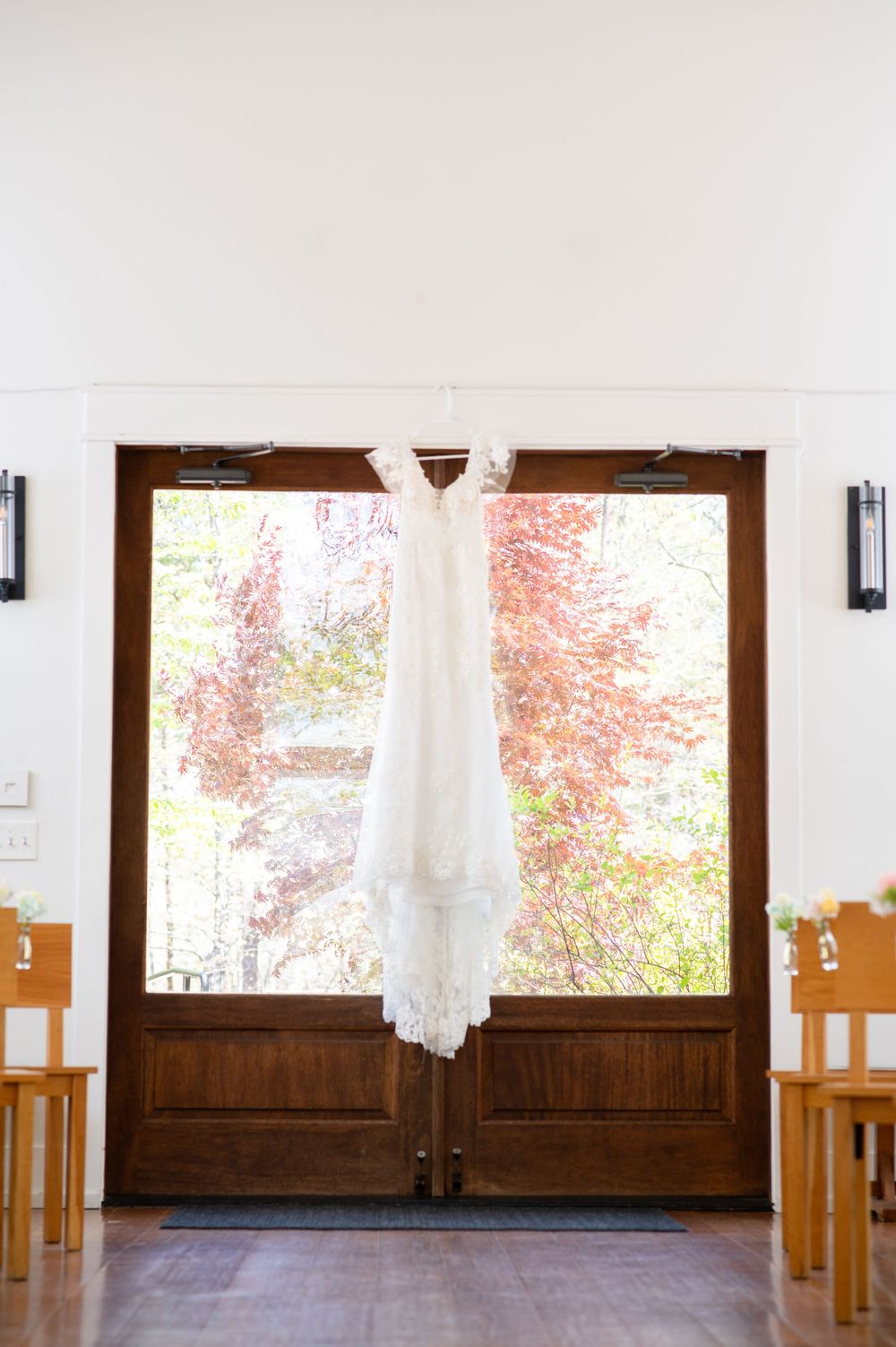 An elegant white lace wedding dress hangs from wooden double doors with bright soring foliage visible through the glass panes. Juliette Chap