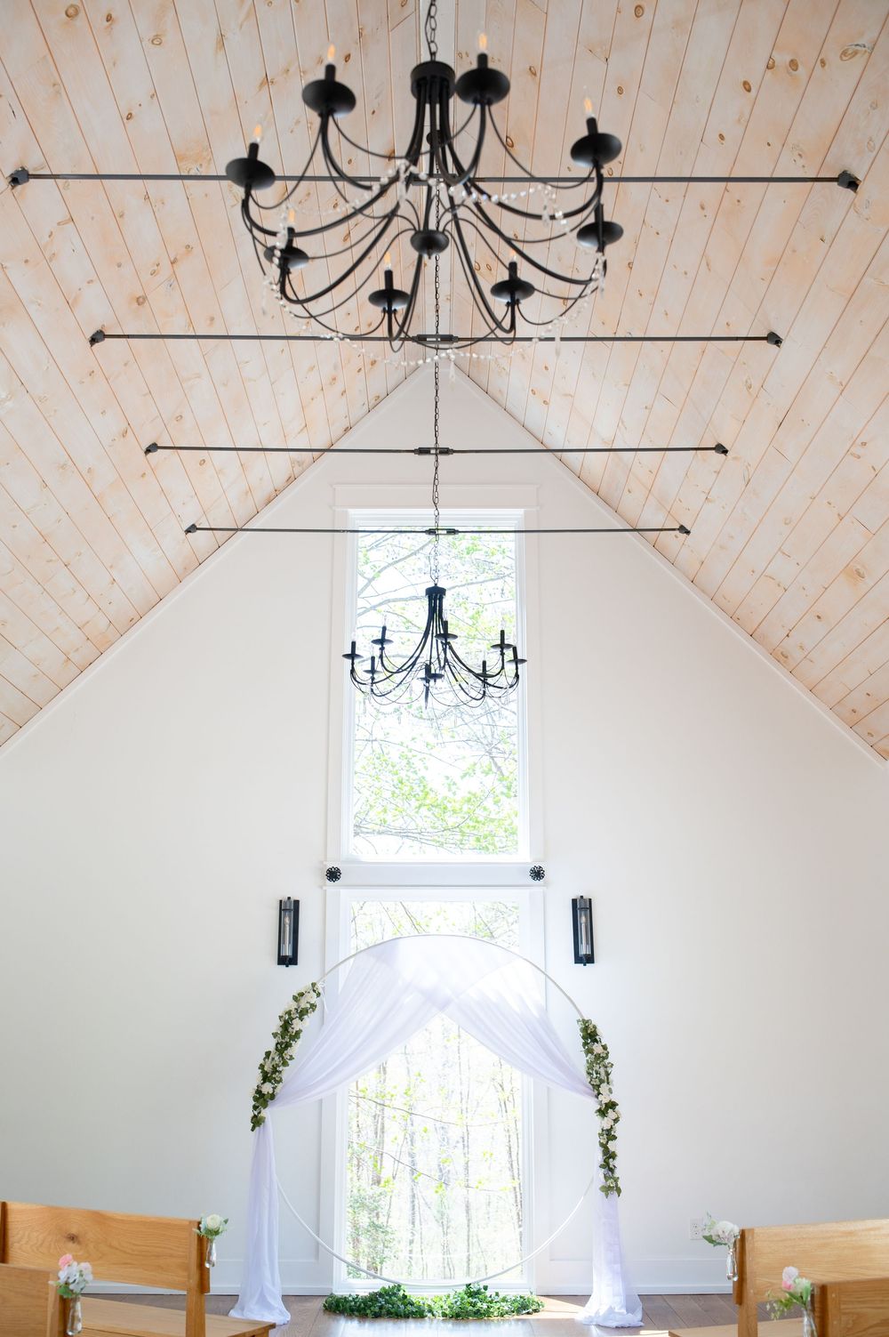 White wedding chapel interior with elegant chandeliers, vaulted wood ceiling, and decorative drapery by the window. Juliette Chapel Wedding