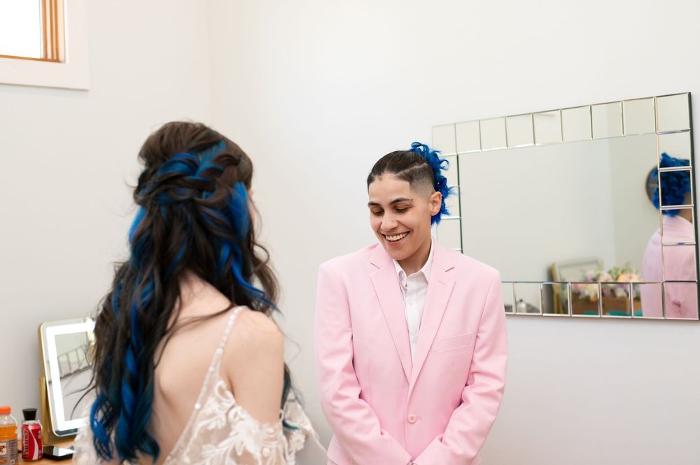 Two people in wedding attire share a joyful moment, with one wearing a pink suit and another in a white dress.