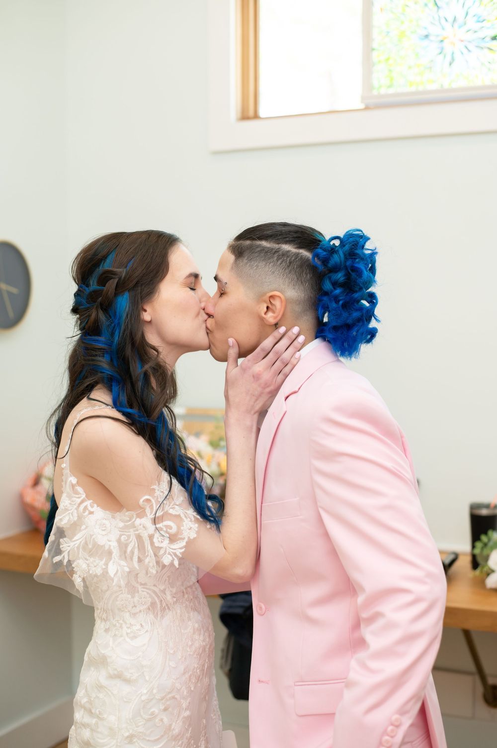 Two people share an intimate kiss at a wedding, with one wearing a white lace dress and the other in a soft pink suit.
