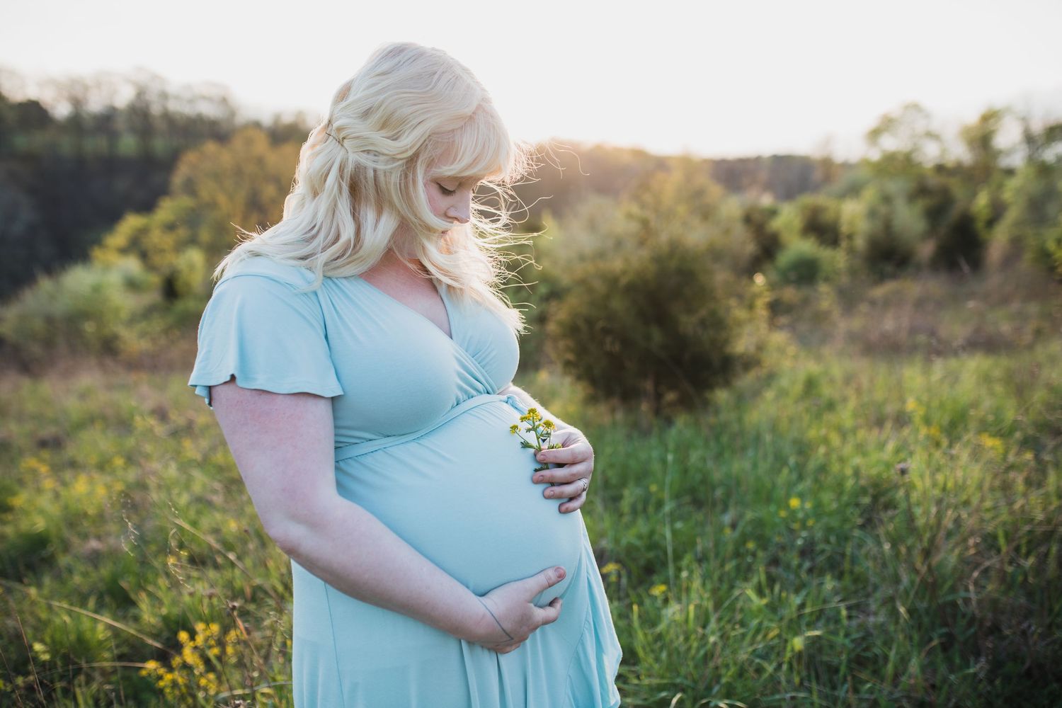 Person in a light blue dress stands in a sunlit meadow during golden hour while cradling their pregnant belly.