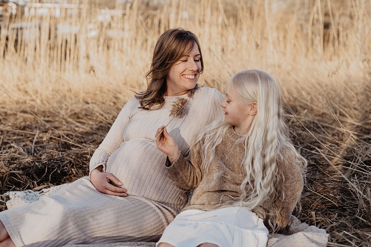 Mot och dotter sitter på en filt vid strandkantens med vass i bakgrunden, flickan håller i ett vasstrå och kittlar sin gravida mamma med.