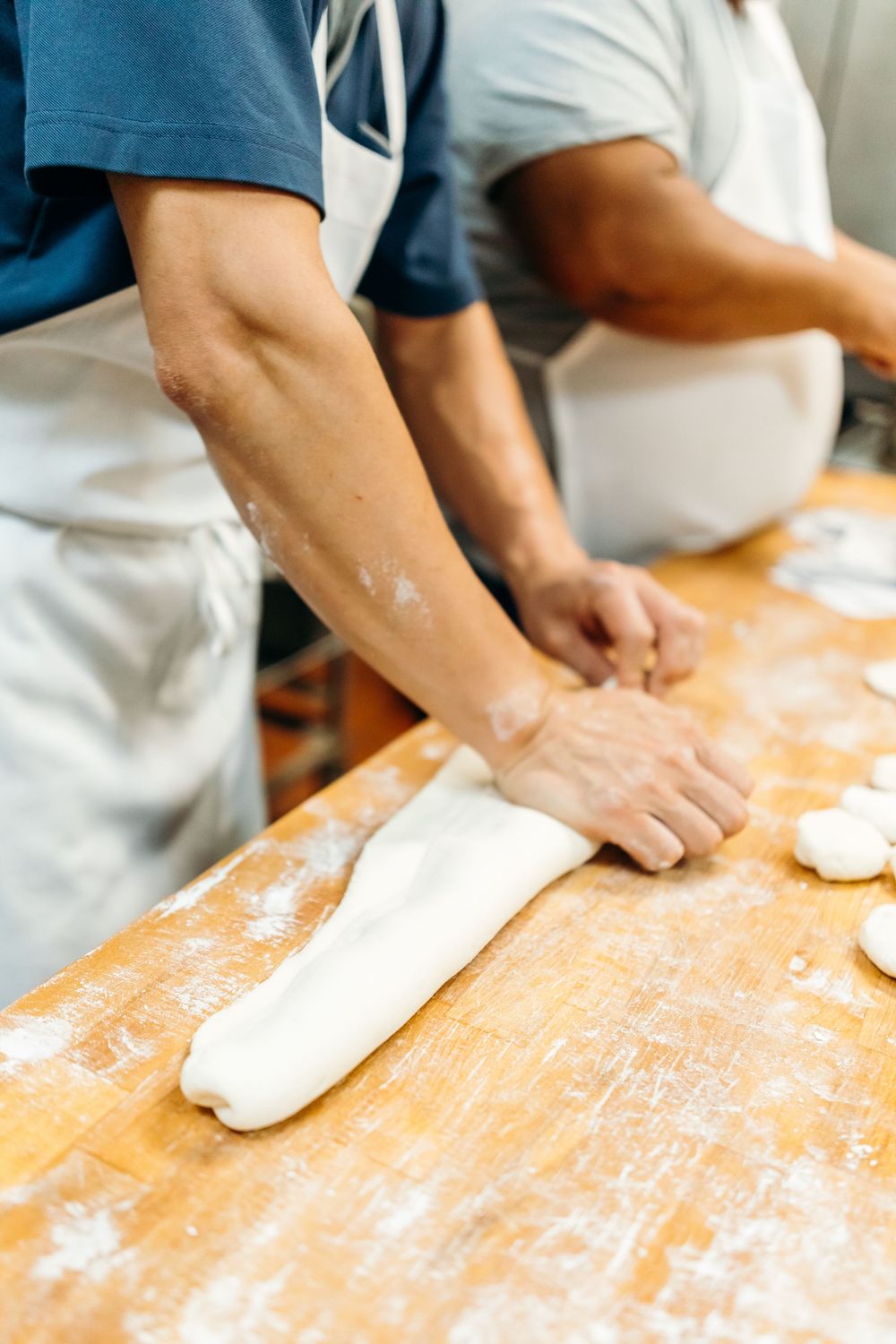 Hands kneading and rolling out fresh dough on a flour-dusted wooden surface in a bakery kitchen.