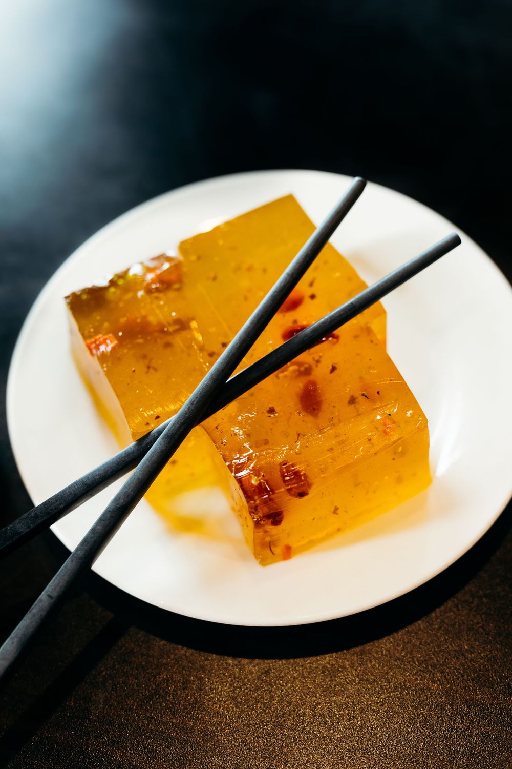 Translucent amber-colored jelly squares with dried chili flakes served on a white plate with black chopsticks.
