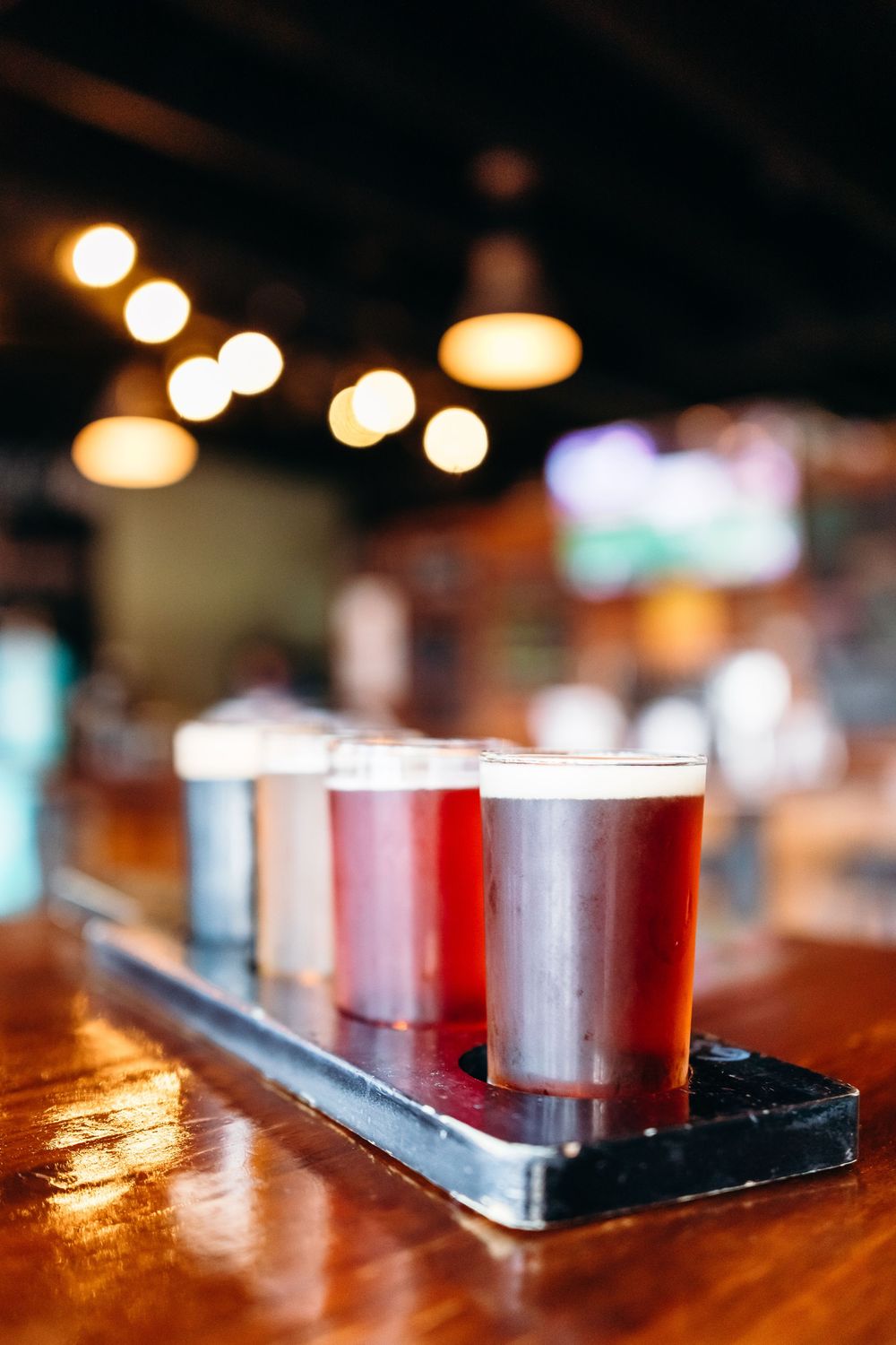 A flight of craft beers in tasting glasses lined up on a wooden bar with bokeh lights in the background.