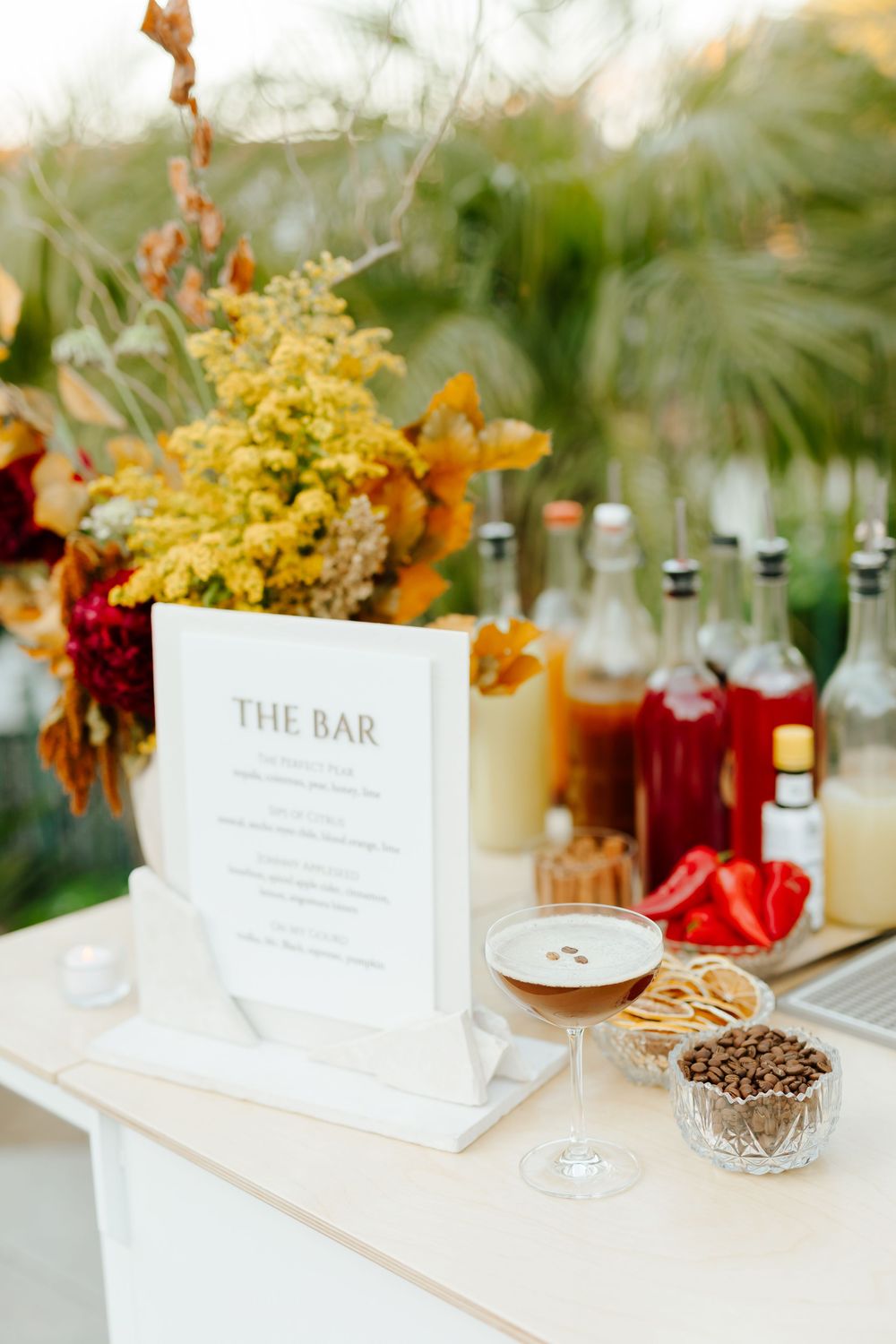 Elegant bar menu display with autumn floral arrangements and assorted drink bottles on a white table at an event.