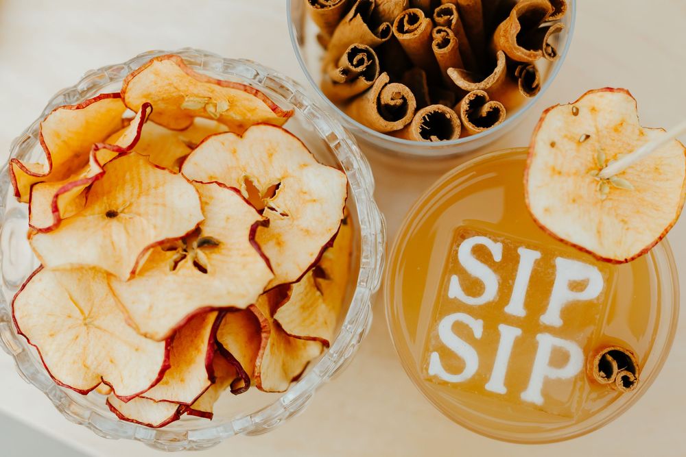 Overhead view of dried apple chips in a bowl next to a beverage with 'SIP SIP' text and cinnamon sticks.