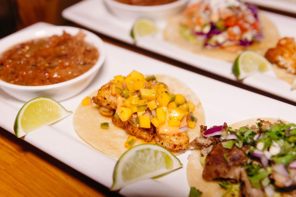 Colorful street style tacos served on white plates with lime wedges and spicy salsa on a wooden table top.