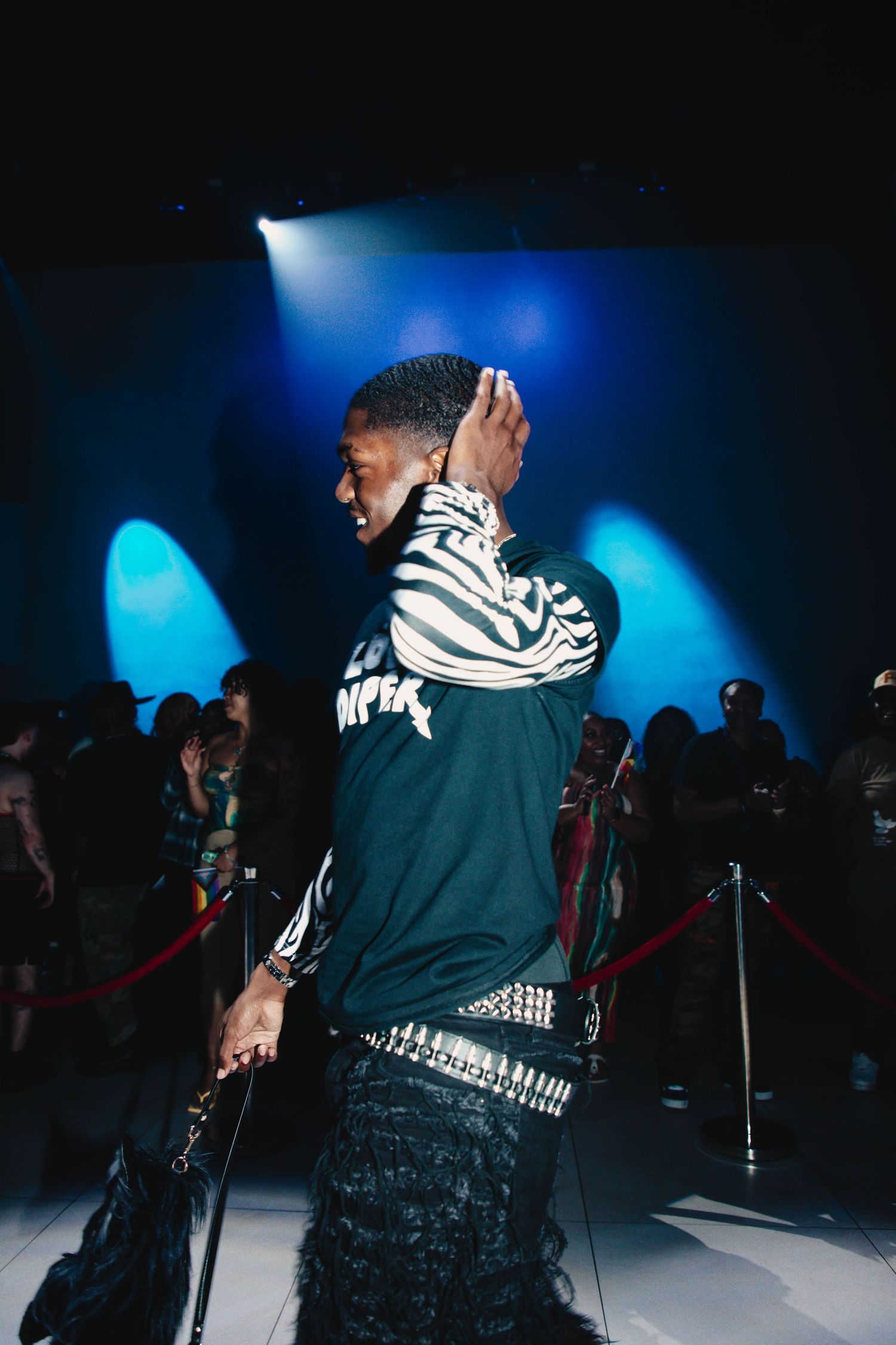 Performer on stage in dark jacket with striped design under dramatic blue lighting and silhouetted crowd behind velvet ropes.