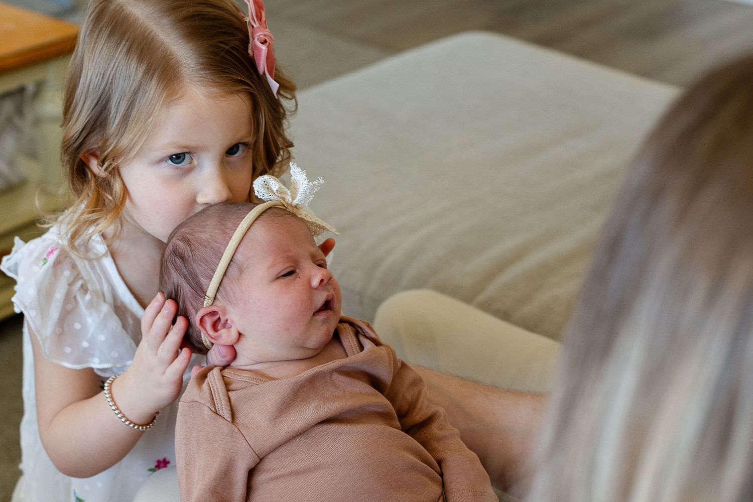 mom holding a newborn with pretty pink bow. Big sis kissing the top of baby's head.