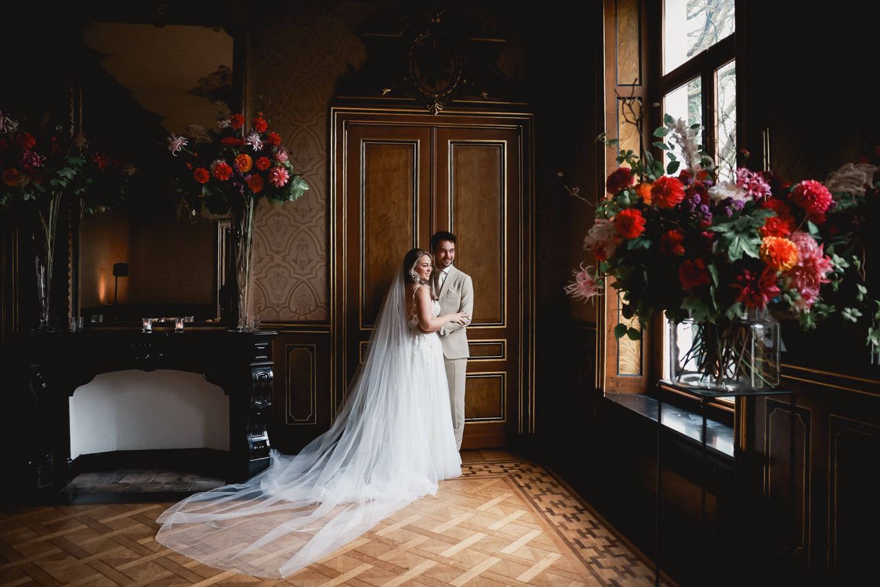 A bride in a flowing white gown stands in a sunlit ballroom surrounded by rich wood paneling and red floral arrangements.