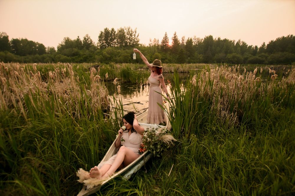 Two people in white dresses relax in a golden meadow at sunset among tall cattails and wild grasses.