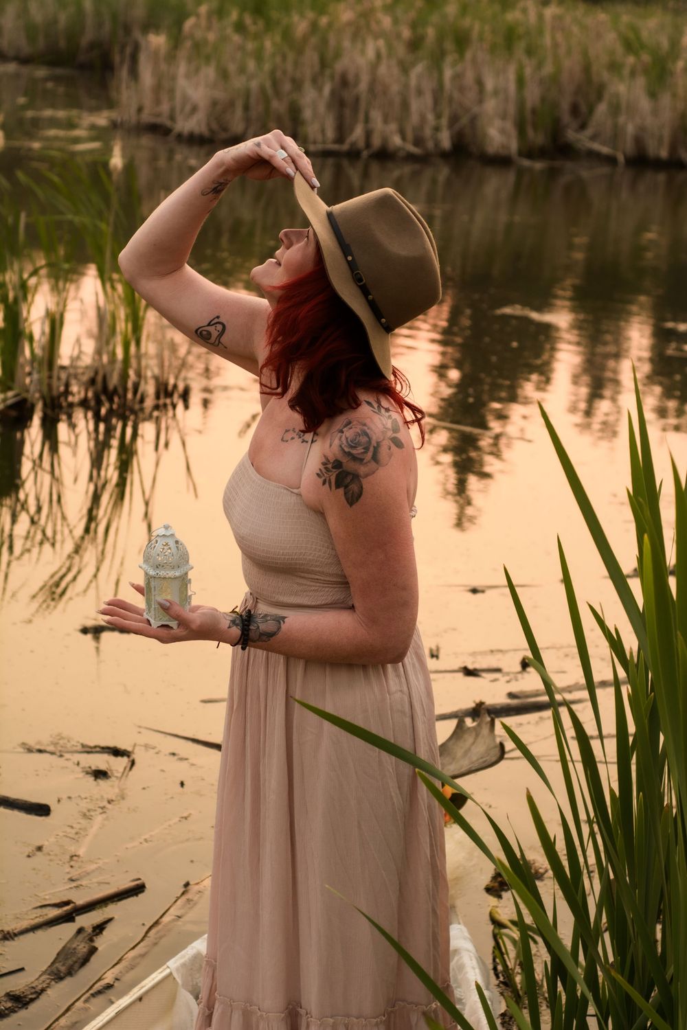Person in beige dress and hat poses by a tranquil pond with reeds at sunset while holding a water bottle.