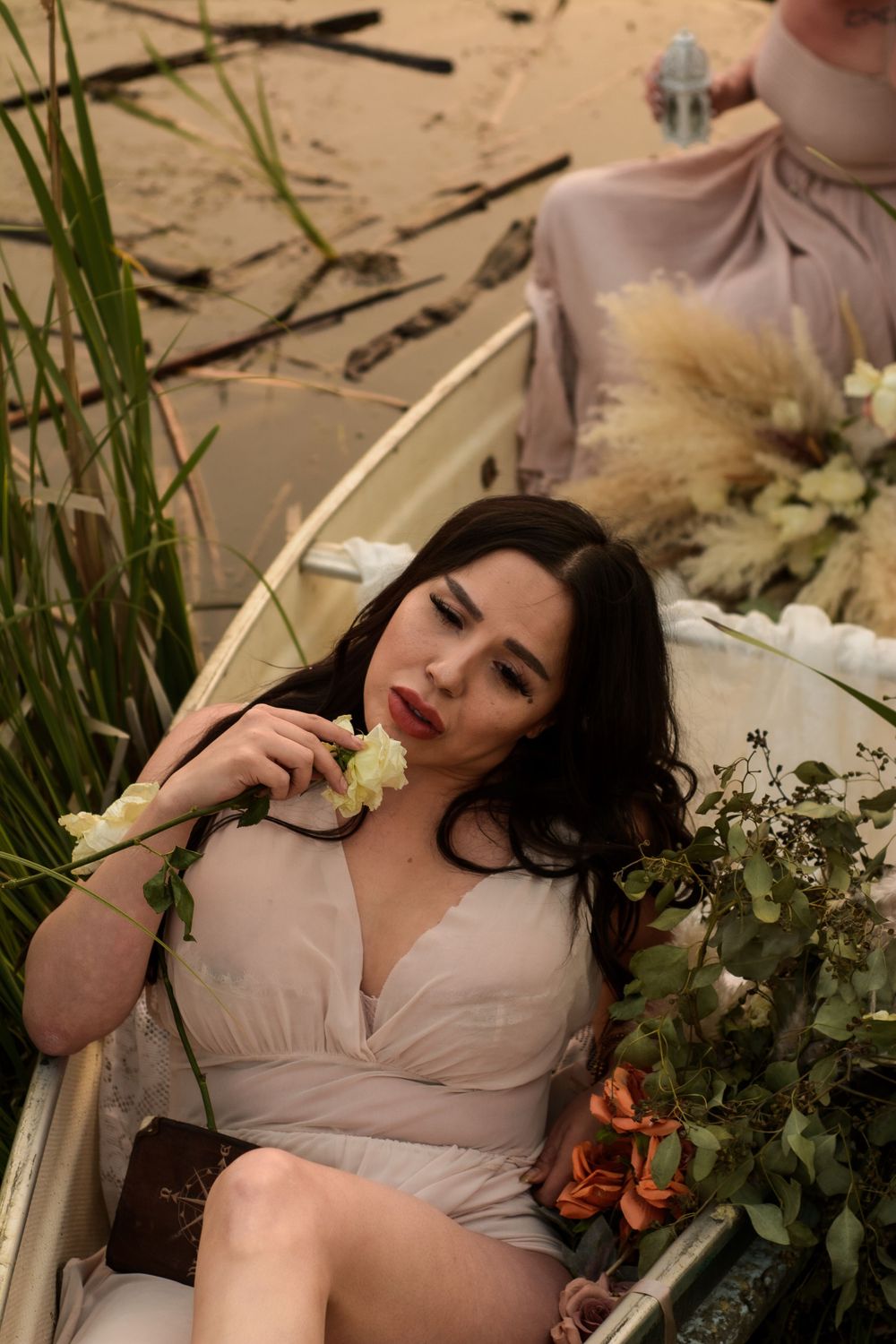 A woman in a blush-colored dress reclines in a rustic rowboat surrounded by flowers and greenery near the water.