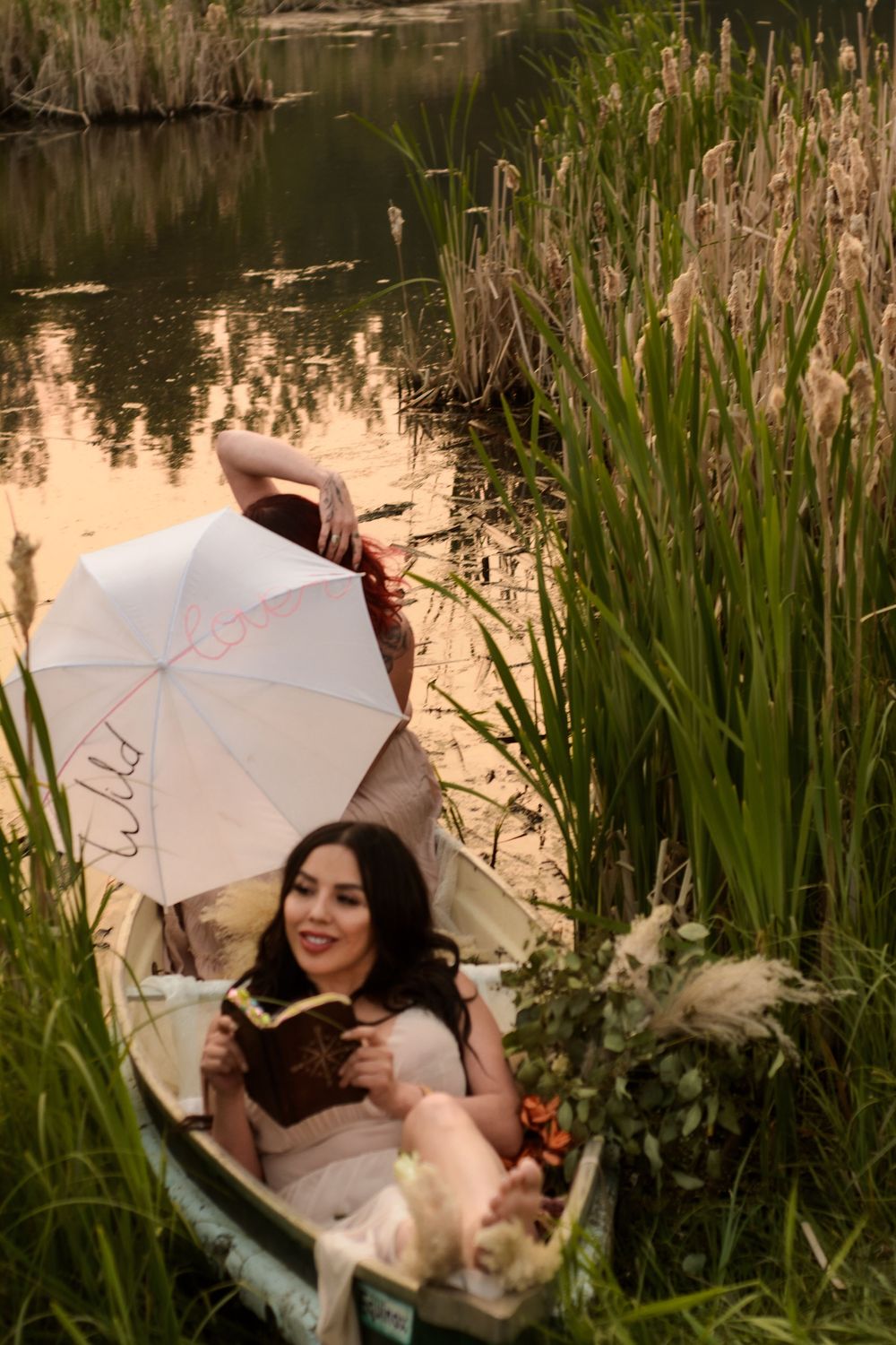 Someone relaxing in a small boat on a serene pond while reading and holding a white parasol among cattails at sunset.
