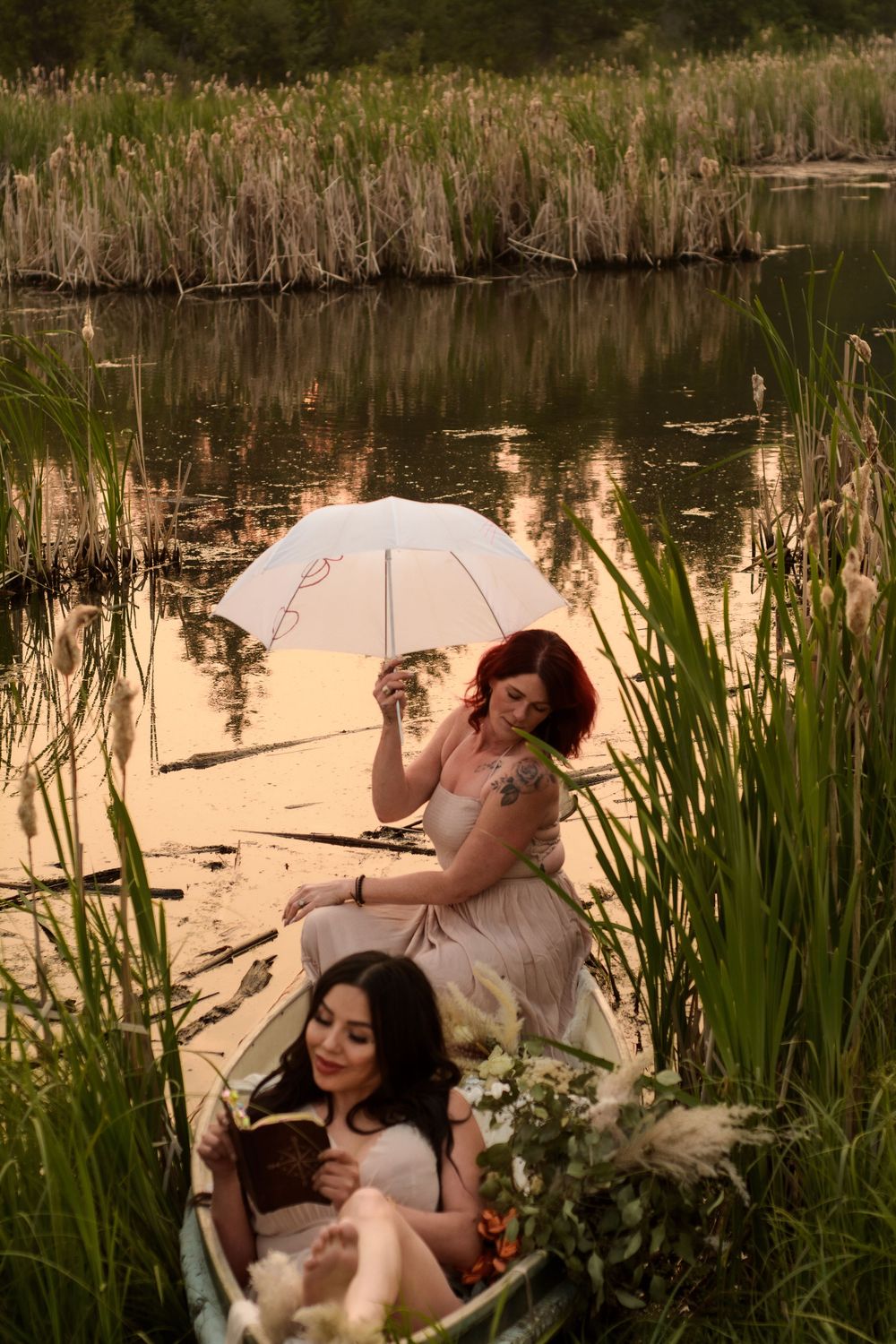Two people relax in a rowboat on a tranquil pond at sunset, with cattails and golden reflections on the water.