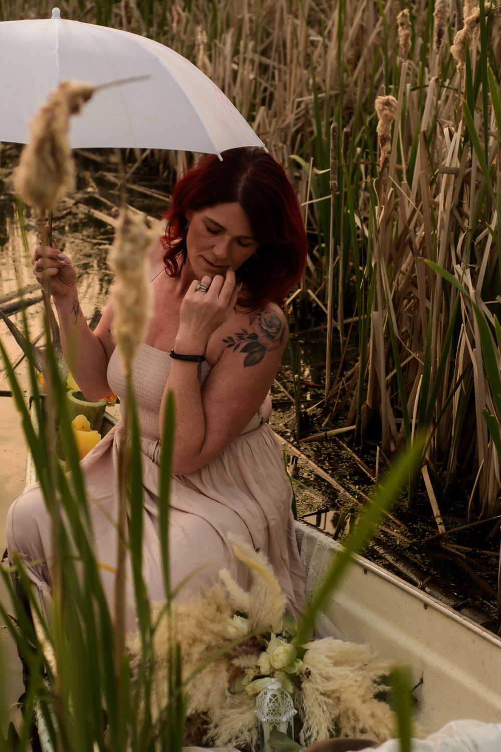A figure in a white dress holds a parasol while sitting among tall cattails and grass in warm natural lighting.