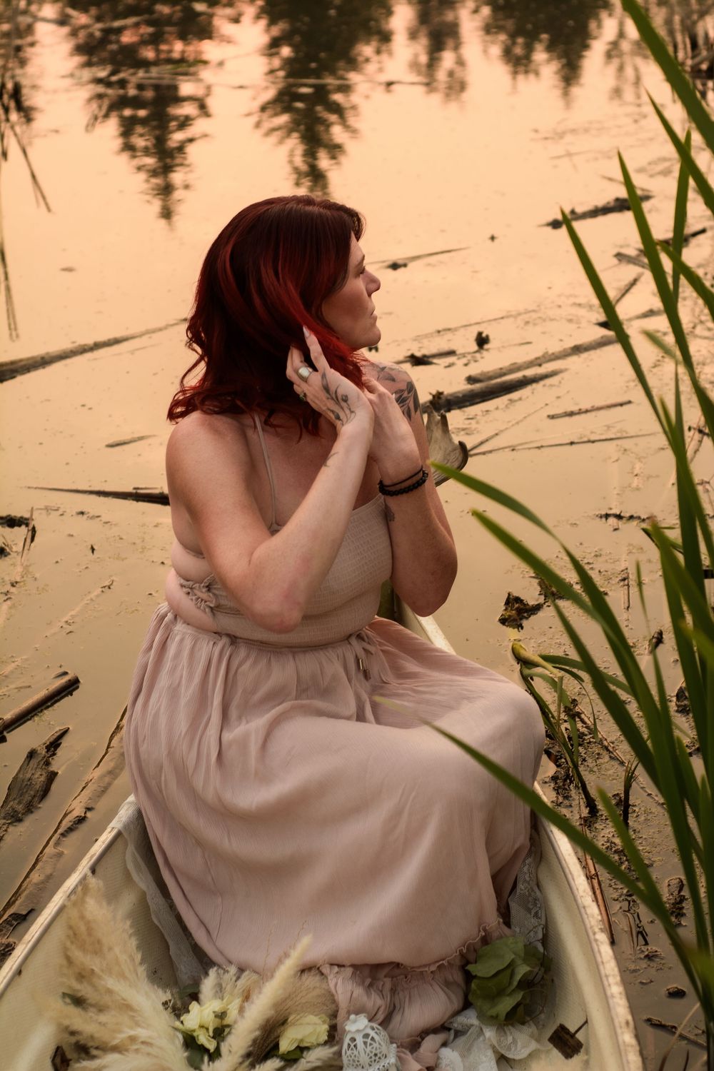 A person in a flowing beige dress sits contemplatively in a wooden boat surrounded by reeds at sunset.