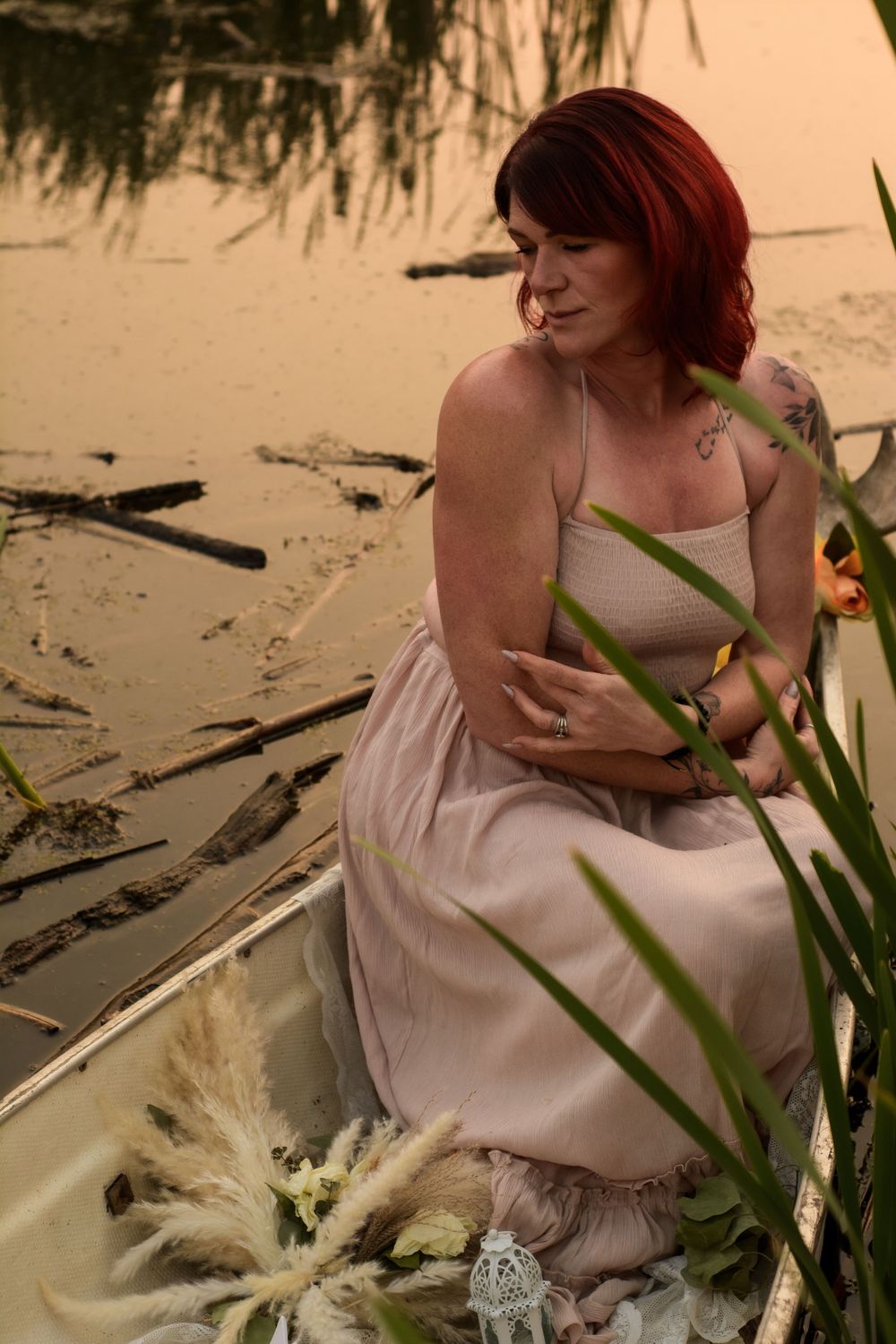 Woman in light dress sits thoughtfully in boat surrounded by cattails and flowers at sunset.
