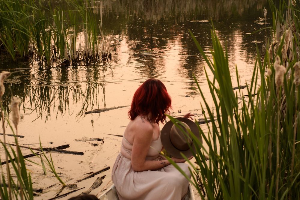A person in a white dress sits by a tranquil pond at sunset surrounded by reeds and marsh grass.