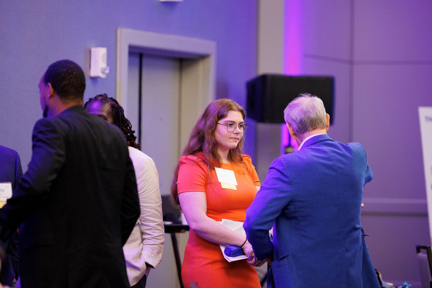 At a networking event with purple lighting, people in professional attire engage in conversation near an elevator.