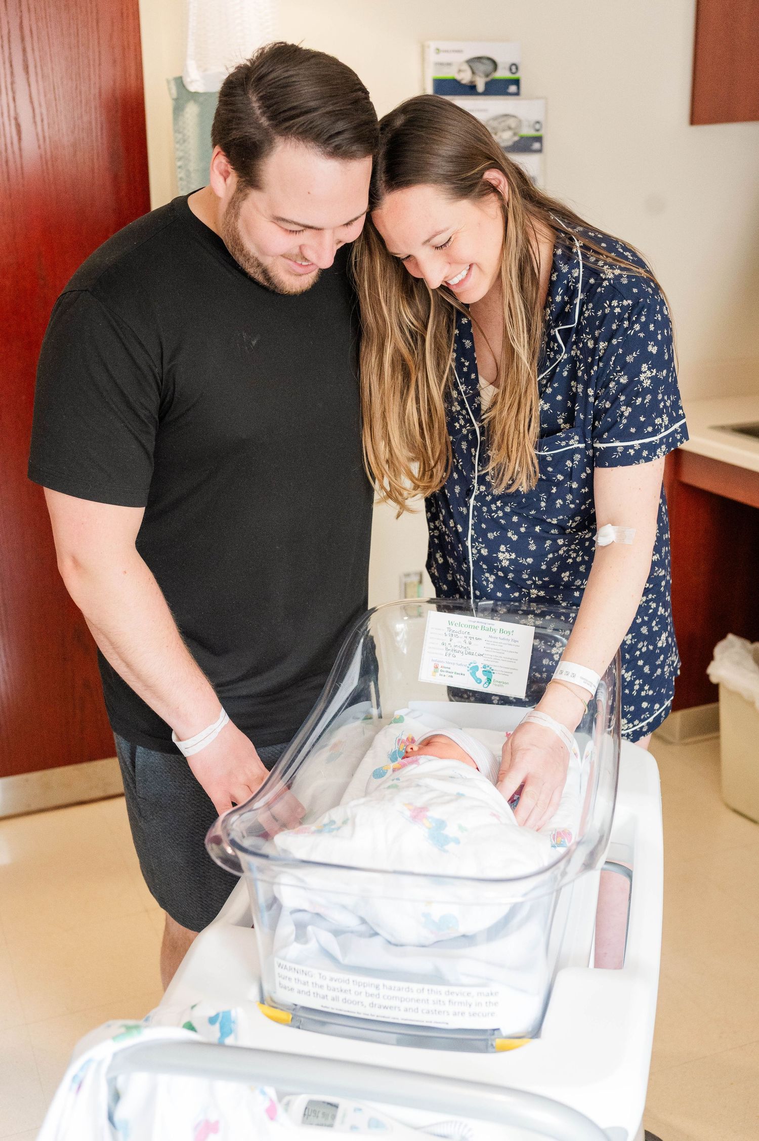 A couple lovingly gazes at a newborn baby sleeping in a hospital bassinet.