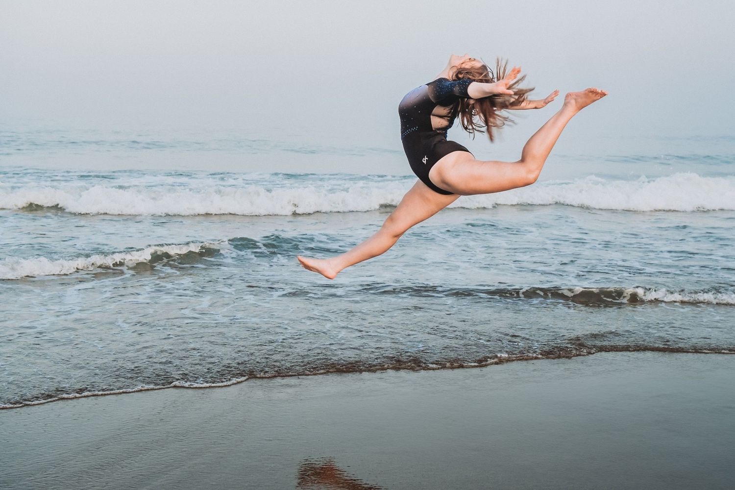 A HS senior who is a gymnast in a black leotard leaps gracefully above the ocean waves at the beach during sunset at Reid State Park.