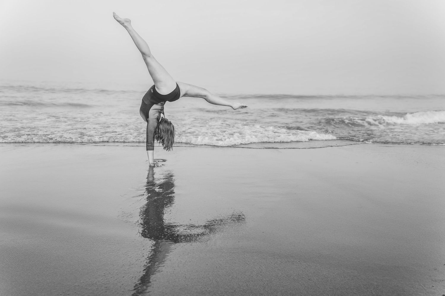A high school senior and gymnast performs a graceful handstand on a reflective beach shoreline at dusk in black and white photography.