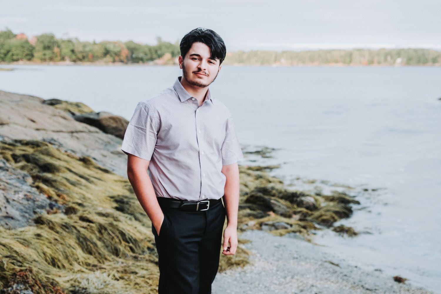 A HS senior stands along a rocky shoreline wearing a light colored shirt and dark pants by a serene waterfront at Wolfe's Neck State Park.