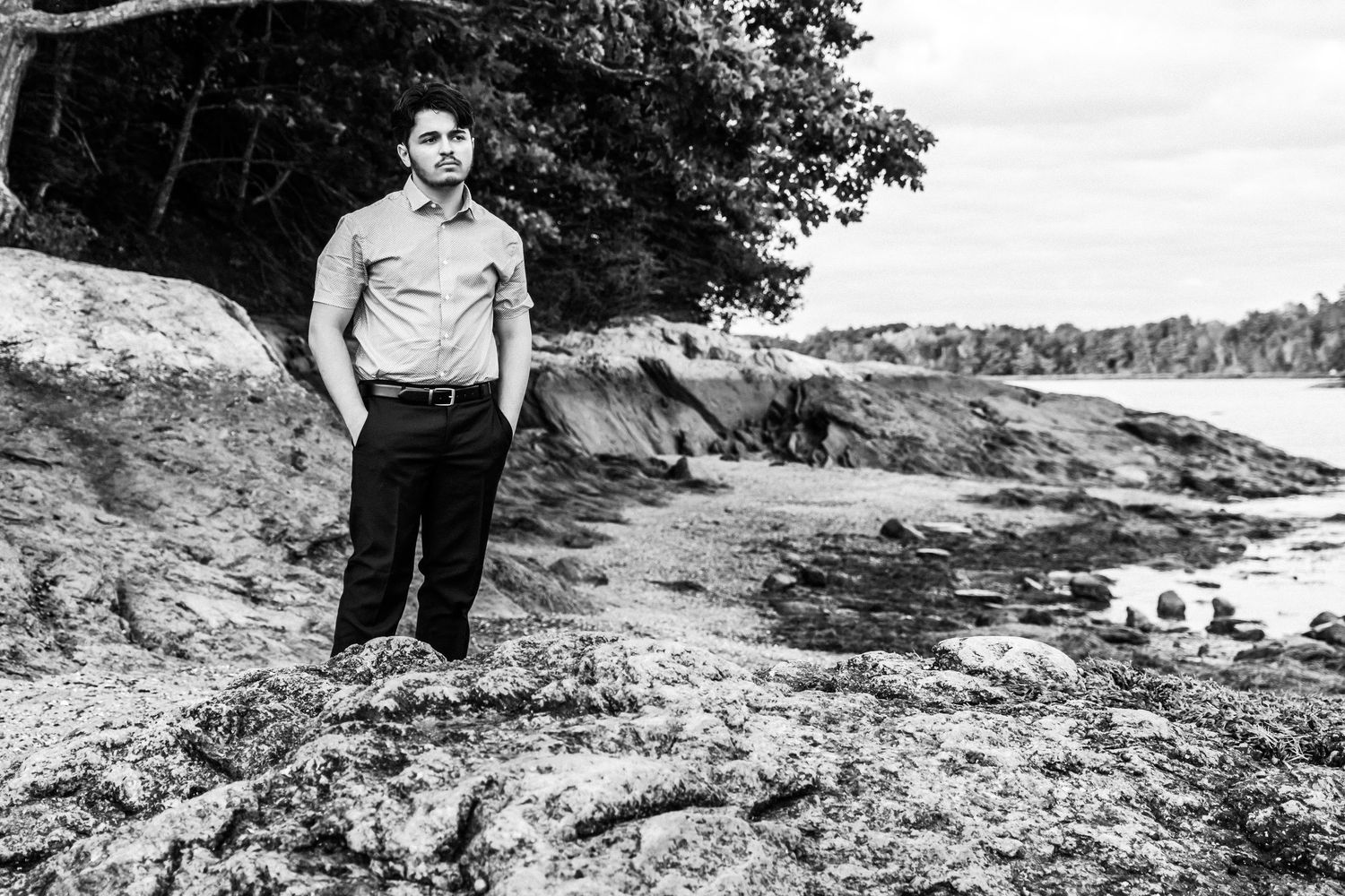 Black and white photograph of a young man who is graduating from HS, standing on rocky coastal terrain with trees and ocean in background.