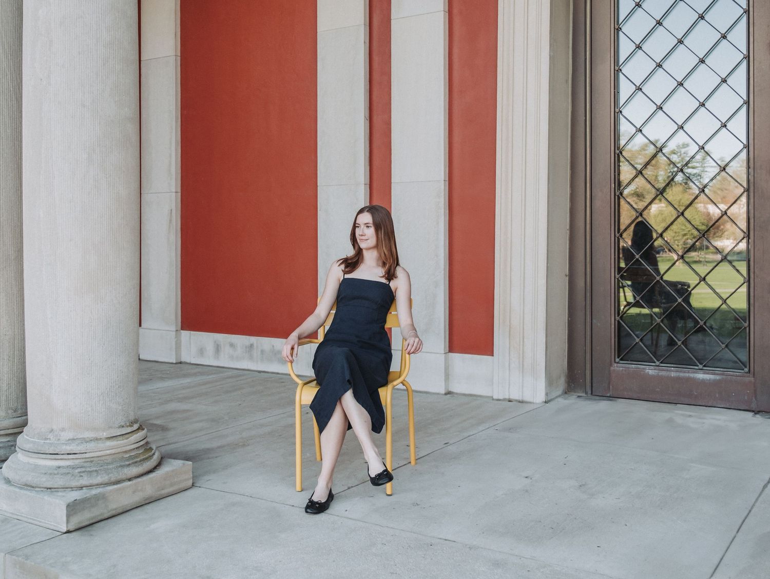 A recent Bowdoin College graduate sits elegantly in a yellow chair against a coral-colored wall with classical columns at the Art Museum.