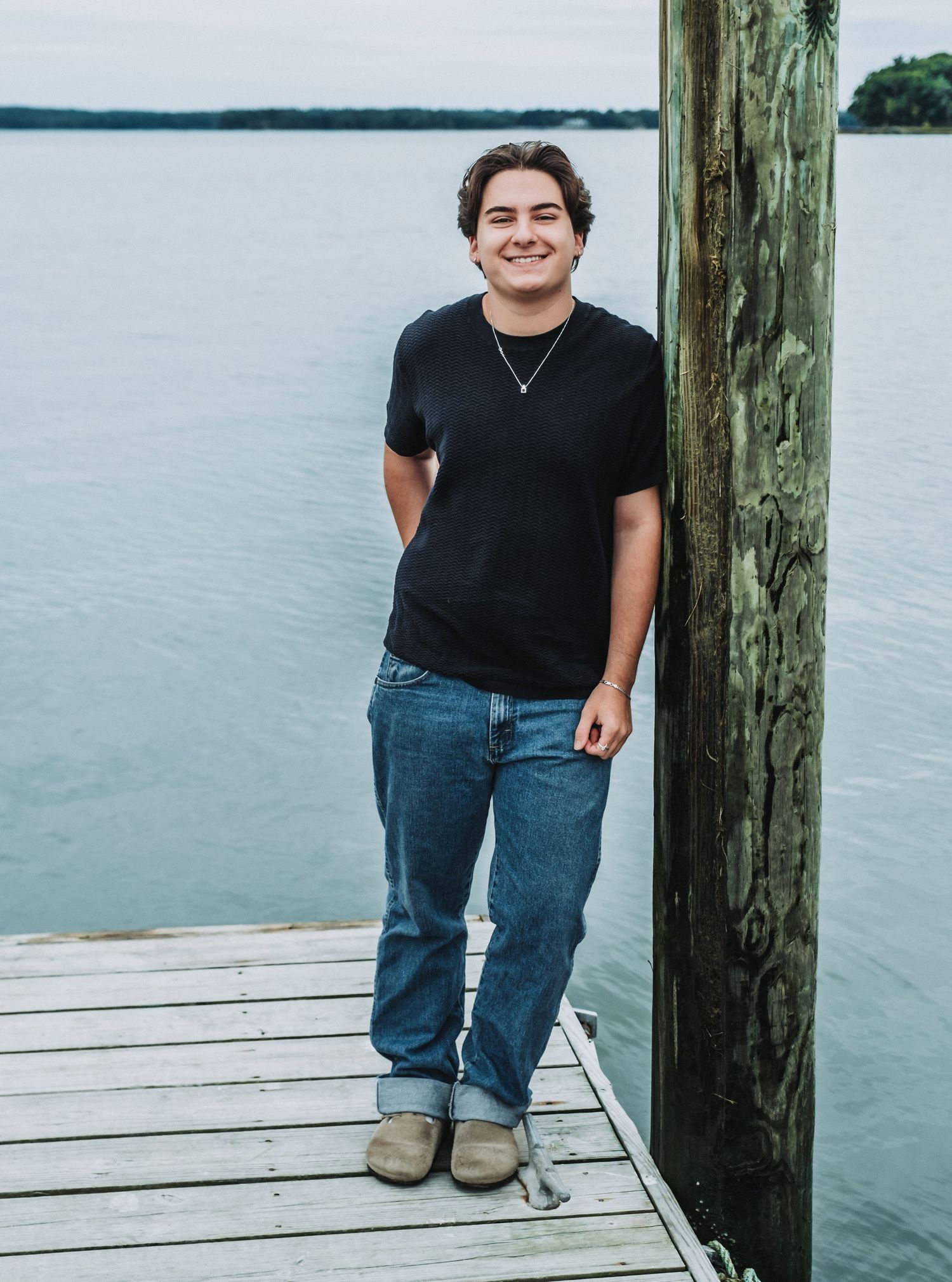 A senior from Freeport High School stands on a wooden dock by a calm water at Winslow Park wearing a black t-shirt and jeans.