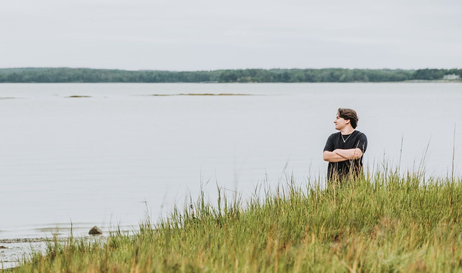 A HS senior stands overlooking a serene coastal landscape with tall grass in the foreground and water stretching to the horizon.